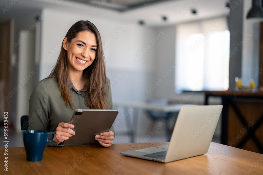 Portrait of smiling fulfilled young woman holding digital tablet device, looking at camera, sitting at desk at home with laptop computer in front of her and cup of coffee. Portrait of smiling fulfilled young woman holding digital tablet device, looking at camera, sitting at desk at home with laptop computer in front of her and cup of coffee.
