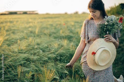 Beautiful woman with wildflowers and straw hat walking in barley field in sunset light. Stylish female relaxing in evening summer countryside and gathering flowers. Atmospheric tranquil moment