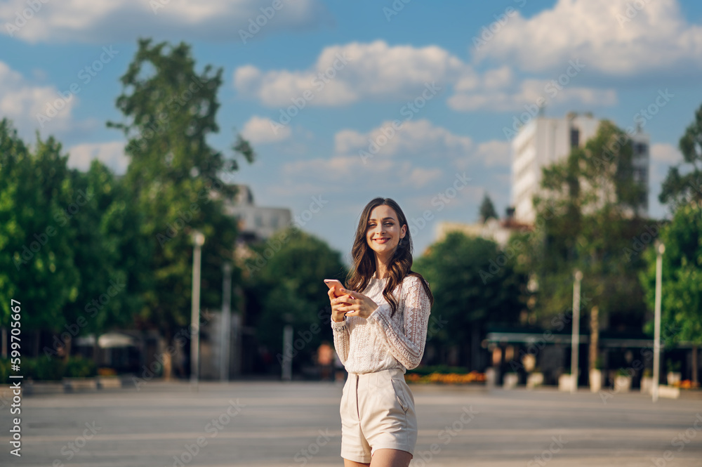 A cheerful young woman in summer style clothes is posing on the city square downtown with her phone and smiling at the camera.