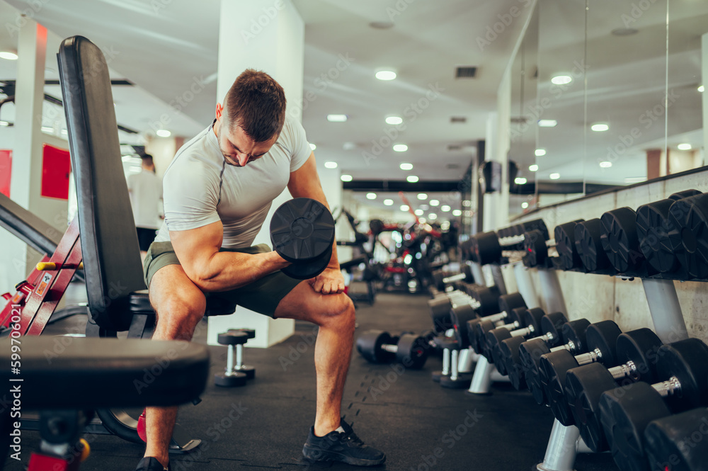 A strong sportsman is sitting in a gym next to the rack with dumbbells and exercising his biceps and triceps. A strong sportsman is sitting in a gym next to the rack with dumbbells and exercising his biceps and triceps.