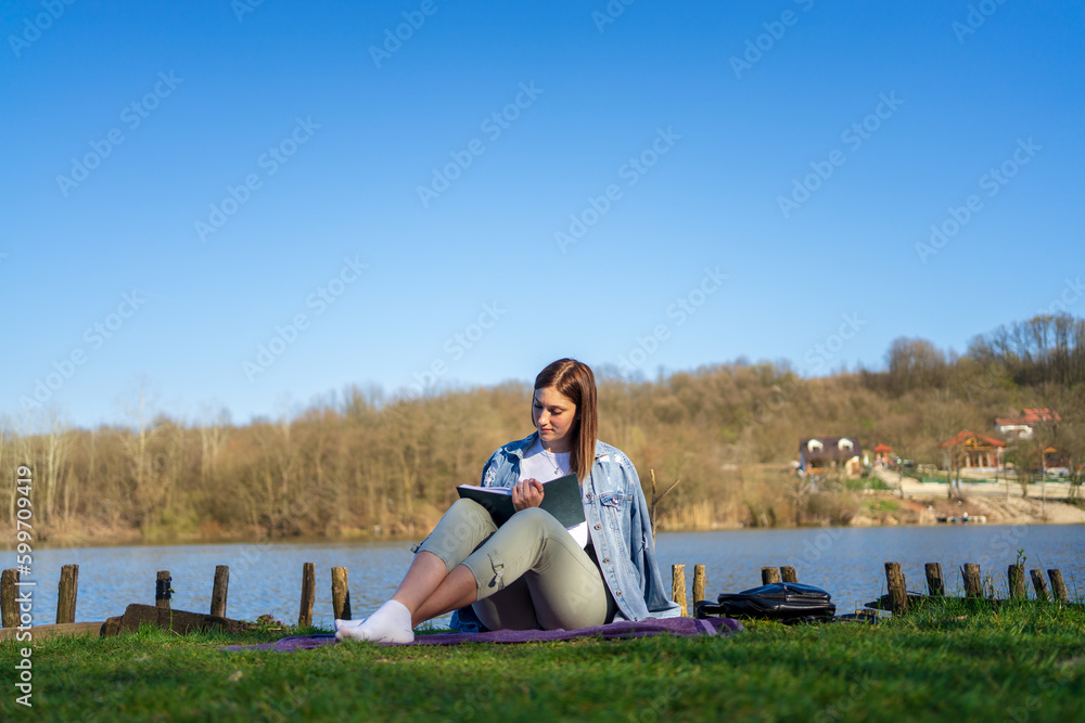 Fototapeta premium Woman at lakeside writing in a notebook. Nature concept. Self reflection.