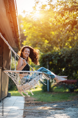 happy young female riding on macrame swing chair near country house outdoors