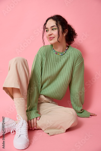 Vertical studio shot of teenage girl sitting on floor in front of a pink background wearing green sweater, beige pants, white sneakers and looking away has positive expression smiling and posing.