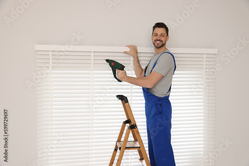 Worker in uniform installing window blind on stepladder indoors