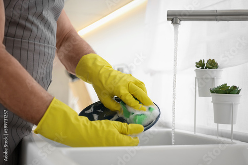 Man in protective gloves washing plate above sink in kitchen, closeup