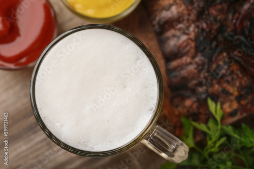 Mug with beer, delicious grilled ribs and sauces on wooden table, closeup