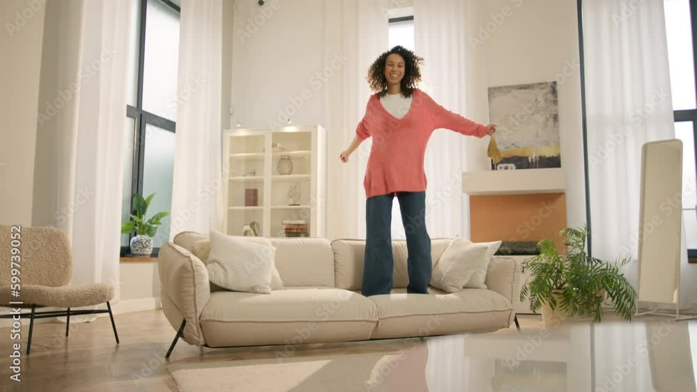 Happy young black woman jumping on sofa, feels overjoyed by celebrating move in day to own home. Mixed race woman laughing enjoying newly rented modern flat or urban apartment. Fashion furniture store Happy young black woman jumping on sofa, feels overjoyed by celebrating move in day to own home. Mixed race woman laughing enjoying newly rented modern flat or urban apartment. Fashion furniture store