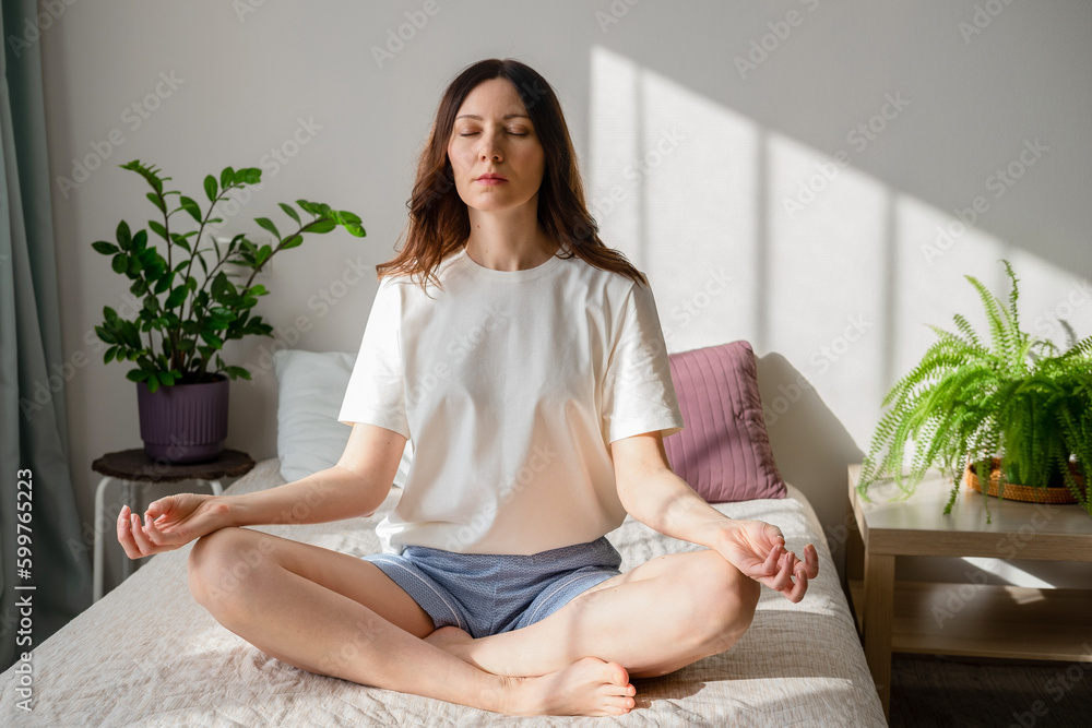 Pretty female sitting on bed in lotus posture with hands on her chest and belly doing pranayama techniques. Doing yoga at home. Self care and positive emotions. Pretty female sitting on bed in lotus posture with hands on her chest and belly doing pranayama techniques. Doing yoga at home. Self care and positive emotions.