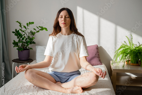 Pretty female sitting on bed in lotus posture with hands on her chest and belly doing pranayama techniques. Doing yoga at home. Self care and positive emotions.