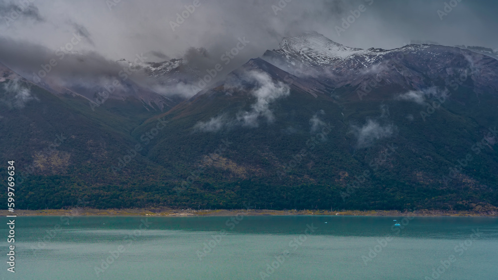Blue fragments of ice are visible on the surface of the turquoise lake. The peaks of the coastal snow-capped mountains are hidden in clouds and fog. Argentina. Lago Argentino