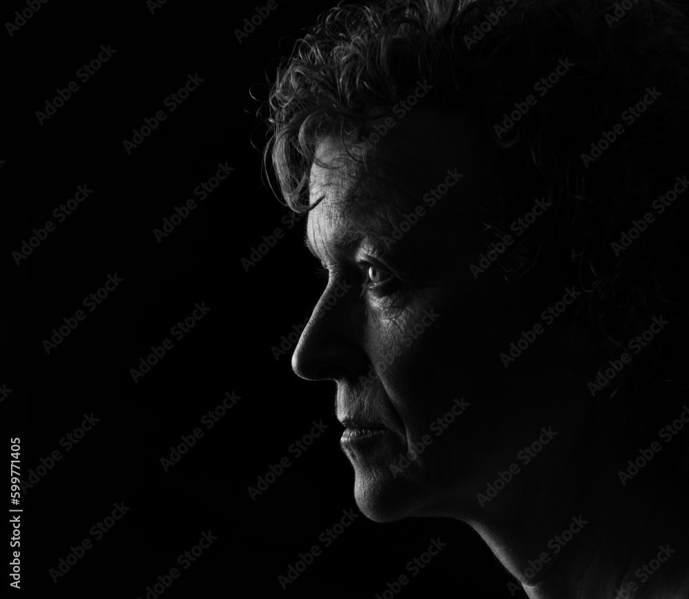 Black and white profile portrait of a middle aged woman with curly hair and a neutral expression, looking to the right.  Low key background, room for copy.