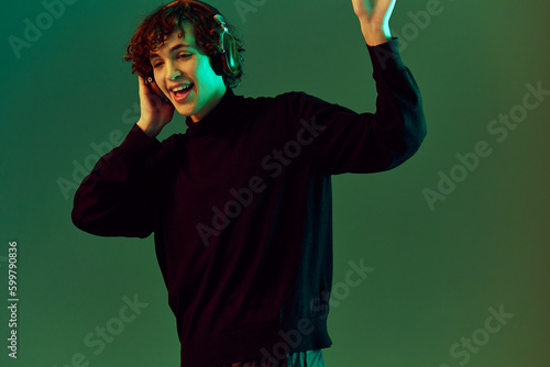 Man wearing headphones listening to music, dancing and singing with his eyes closed, DJ happiness and smile, hipster teen lifestyle, portrait green background mixed neon light, copy space © SHOTPRIME STUDIO