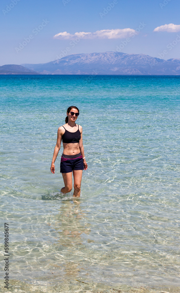 Young woman in black swimsuit and sunglasses walking in sea water, vertical 