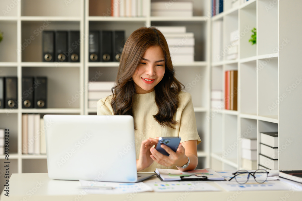 Satisfied asian female financial advisor chatting with client online on smart phone and using laptop at workstation Satisfied asian female financial advisor chatting with client online on smart phone and using laptop at workstation