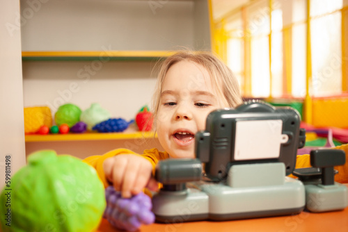 child in kindergarten, playroom, playing with toys, learning. Plays in store, vegetables and fruits, cash register and scales. Funny.