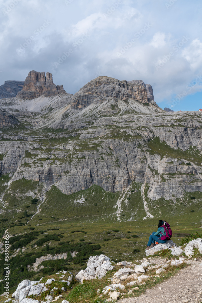 A girl, a tourist in blue clothes with a crimson backpack, sits with her back on a stone and looks at the majestic Dolomite mountains in Italy. Mountain View