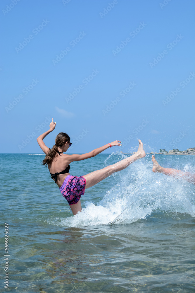 Cheerful smiling girl in sunglasses and bikini splashes in the sea against the blue sky
