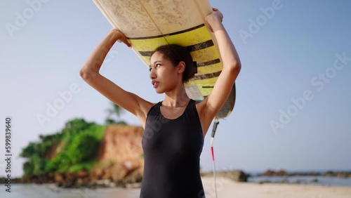 African american woman walking with surfboard on her head on ocean beach. Black female surfer posing with surf board. Pretty multiethnic girl goes on surfing session at tropical location at sunrise.