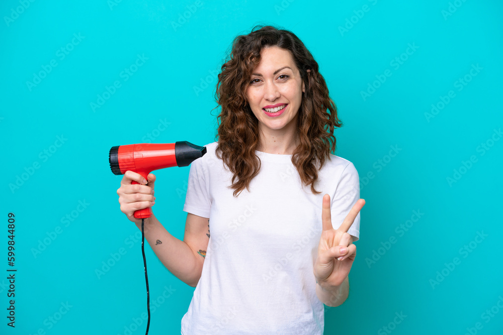 Young caucasian woman holding a hairdryer isolated on blue background smiling and showing victory sign Young caucasian woman holding a hairdryer isolated on blue background smiling and showing victory sign
