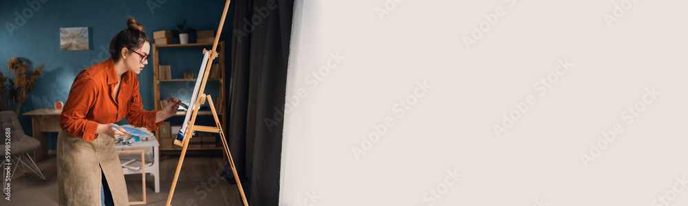 Obraz premium Brazilian woman artist works in concentration. Side view portrait of a creative lady holding paintings on a palette behind an easel in an art studio near a large window. Banner.
