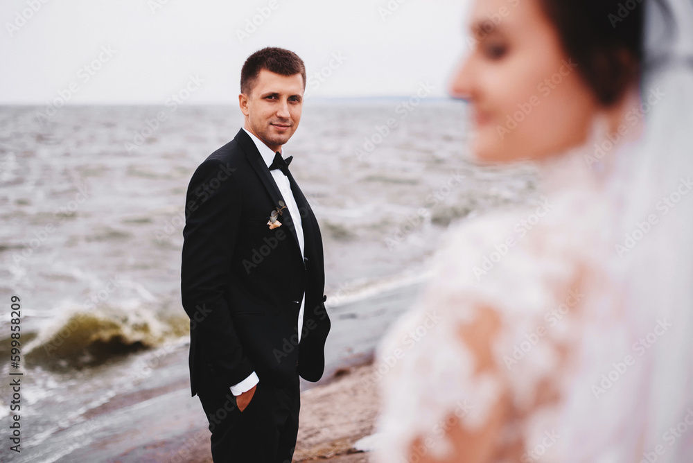 Groom in a black suit with a black bow tie look at beautiful bride in a white long dress near the river. Wedding portrait of newlyweds. Groom in a black suit with a black bow tie look at beautiful bride in a white long dress near the river. Wedding portrait of newlyweds.