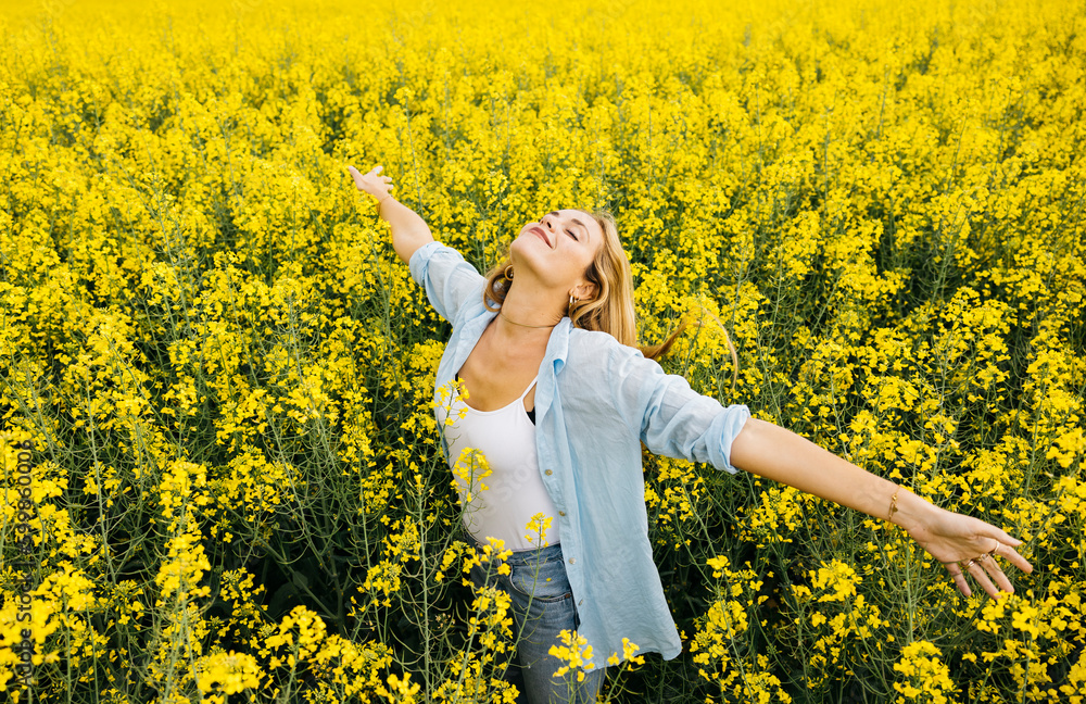 Fototapeta premium Beautiful young blonde cheerful woman, with outstretched arms, amidst a field of blooming yellow rapeseed flowers