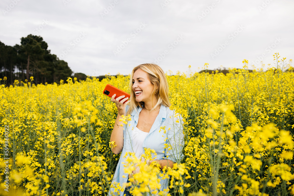 Fototapeta premium Beautiful young blonde cheerful woman, sending a voice message using a red phone, amidst a field of blooming yellow rapeseed flowers