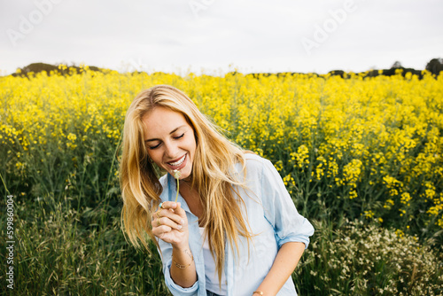 Beautiful young blonde cheerful woman, blowing dandelion seeds amidst a field of blooming yellow rapeseed flowers