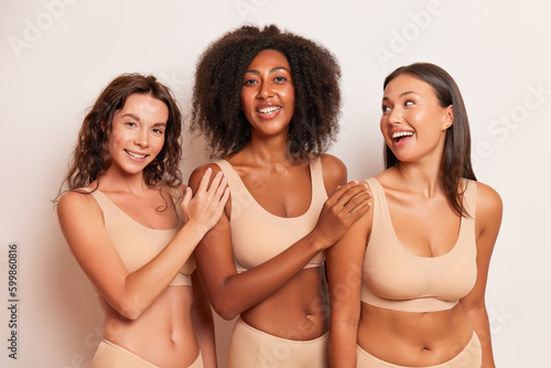 Smiling women with different skin colour in sport underwear pose on white background put hands on shoulder of friend next to, comfort underclothes concept, copy space © South House Studio