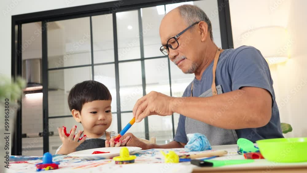 Cheerful Asian Japanese Senior man and little child boy in apron Enjoying painting art at home together. Happy Grandfather coloring on Grandchild hands. Positive emotion. Laughing Cheerful Asian Japanese Senior man and little child boy in apron Enjoying painting art at home together. Happy Grandfather coloring on Grandchild hands. Positive emotion. Laughing