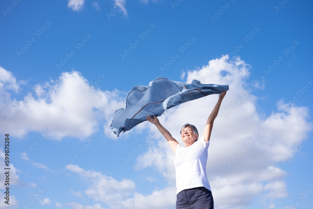 Cheerful senior woman standing outdoors with outstretched arms waving scarf in the blue sky enjoying freedom and vacation Cheerful senior woman standing outdoors with outstretched arms waving scarf in the blue sky enjoying freedom and vacation