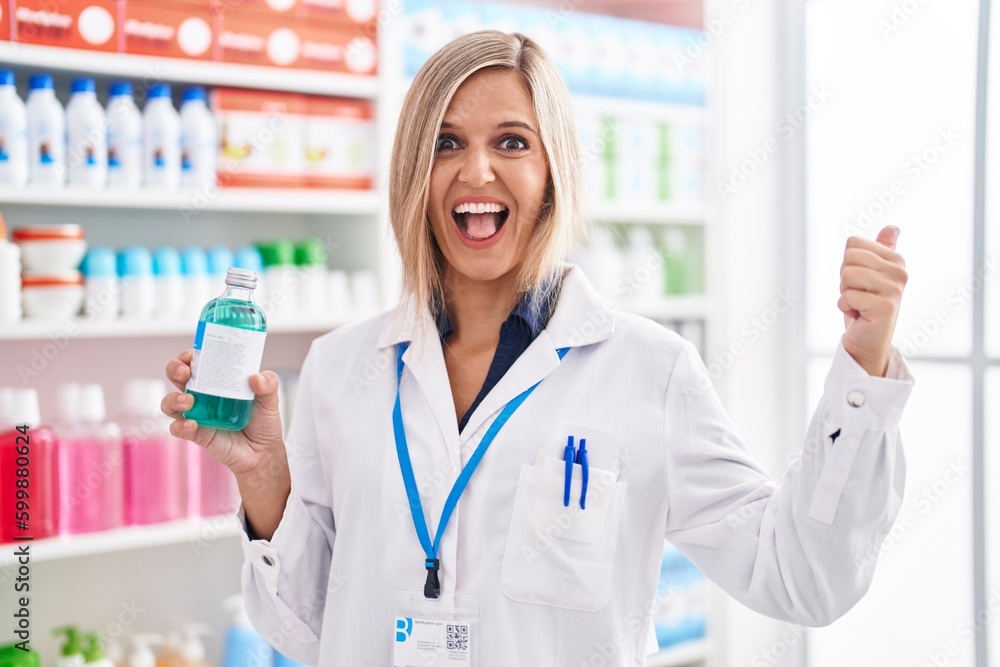 Blonde woman working at pharmacy drugstore holding syrup pointing thumb up to the side smiling happy with open mouth