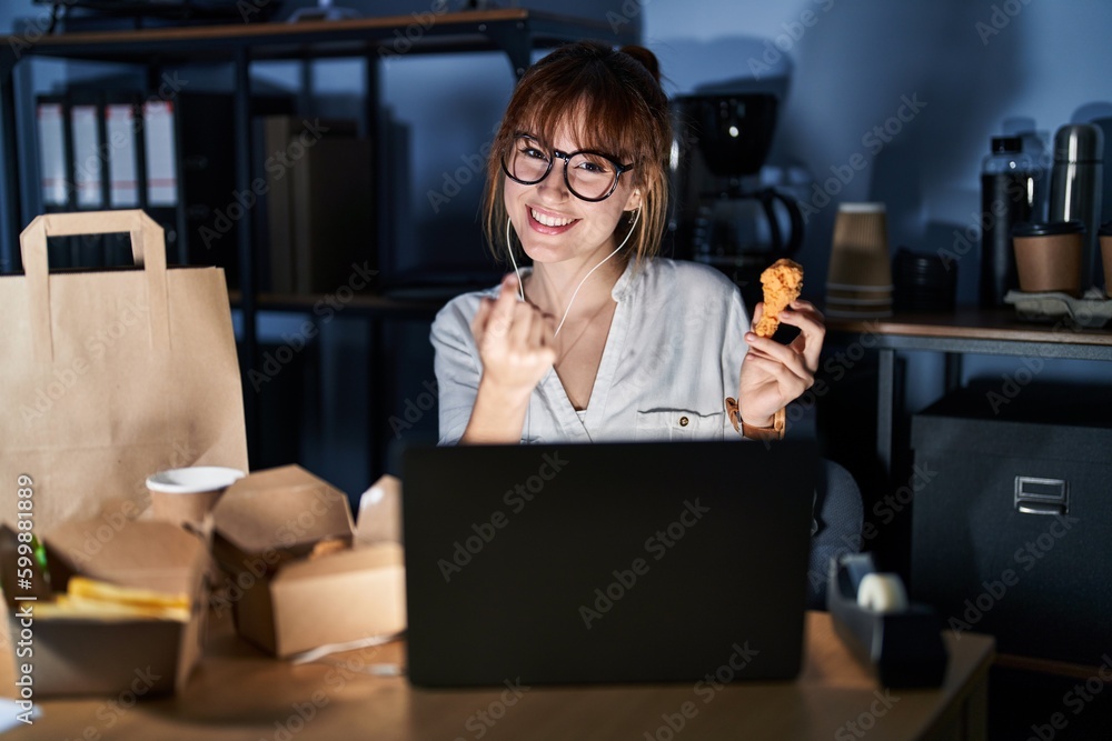 Obraz premium Young beautiful woman working using computer laptop and eating delivery food beckoning come here gesture with hand inviting welcoming happy and smiling