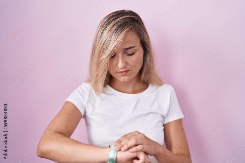 Young blonde woman standing over pink background checking the time on wrist watch, relaxed and confident Young blonde woman standing over pink background checking the time on wrist watch, relaxed and confident
