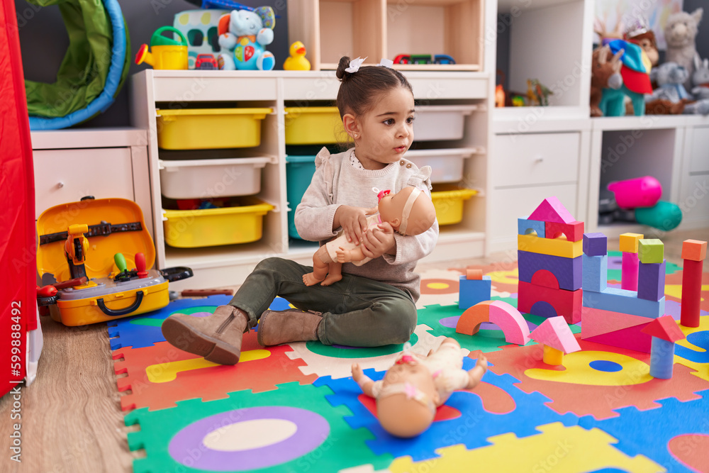 Adorable hispanic girl sitting on floor playing with baby doll at kindergarten Adorable hispanic girl sitting on floor playing with baby doll at kindergarten
