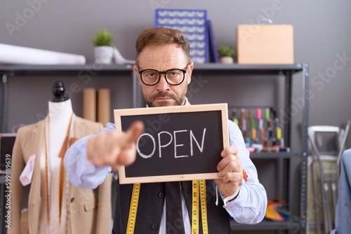 Middle age man with beard dressmaker designer holding open sign pointing with finger to the camera and to you, confident gesture looking serious