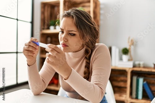 Young beautiful hispanic woman holding pregnancy test sitting on table at home