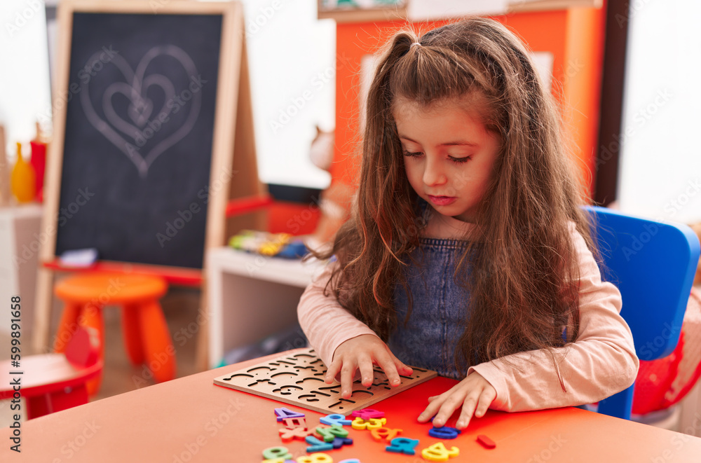 Fototapeta premium Adorable hispanic girl playing with maths puzzle game sitting on table at kindergarten