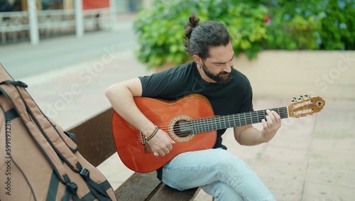 Young hispanic man musician playing classical guitar sitting on bench at park