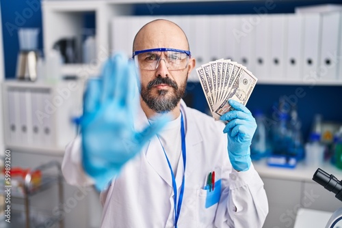 Young hispanic man working at scientist laboratory holding money with open hand doing stop sign with serious and confident expression, defense gesture