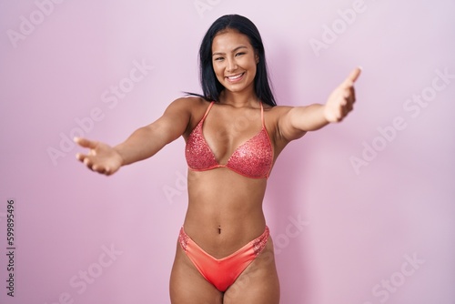 Hispanic woman wearing bikini looking at the camera smiling with open arms for hug. cheerful expression embracing happiness.