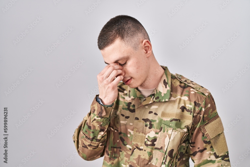Young man wearing camouflage army uniform tired rubbing nose and eyes feeling fatigue and headache. stress and frustration concept.