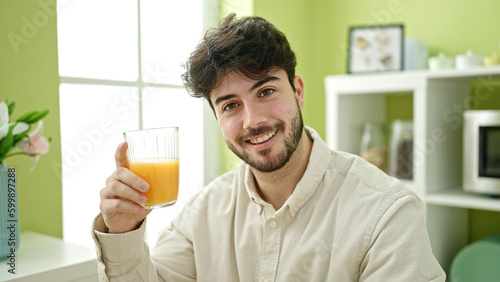 Young hispanic man holding glass of orange juice at dinning room