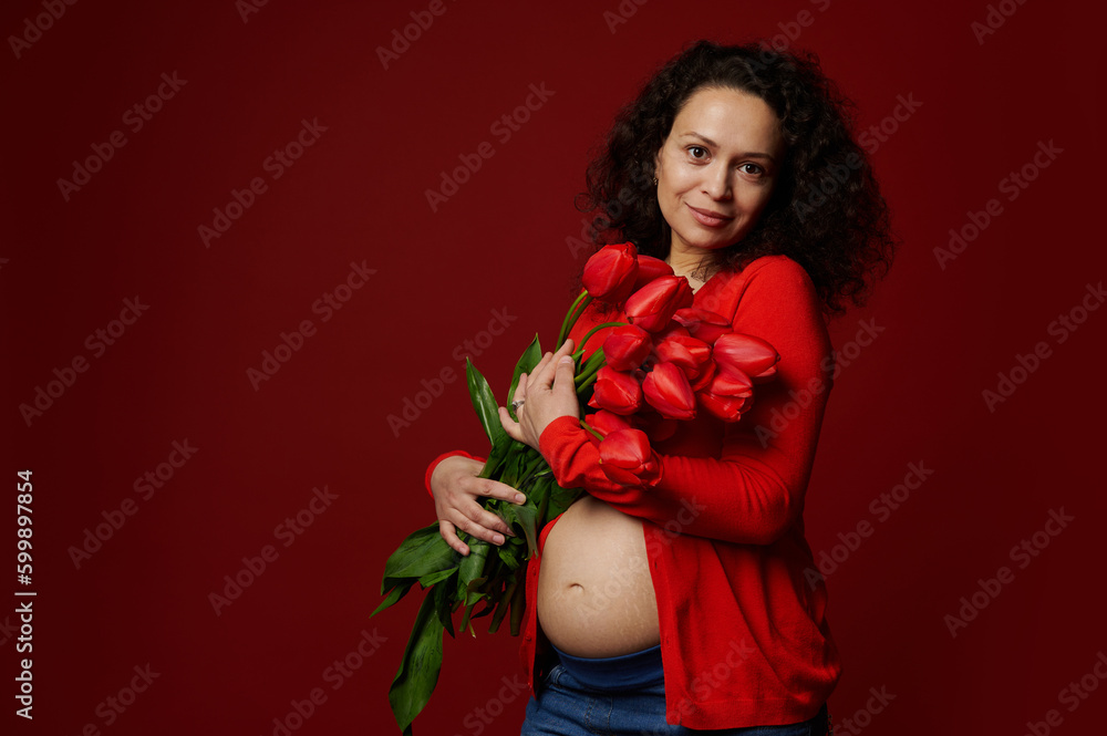 Multi ethnic beautiful pregnant woman, wearing red shirt, holding a cute bouquet of tulips, posing bare belly on isolated color background. Expectant mother. Maternity. Happy Women's and Mother's Day Multi ethnic beautiful pregnant woman, wearing red shirt, holding a cute bouquet of tulips, posing bare belly on isolated color background. Expectant mother. Maternity. Happy Women's and Mother's Day