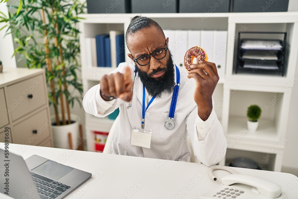 African american man working at dietitian clinic holding doughnut pointing with finger to the camera and to you, confident gesture looking serious