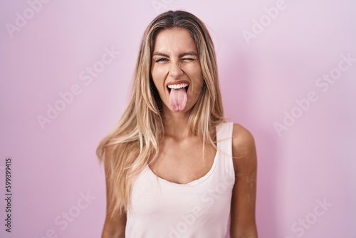 Obraz na plátně Young blonde woman standing over pink background sticking tongue out happy with funny expression