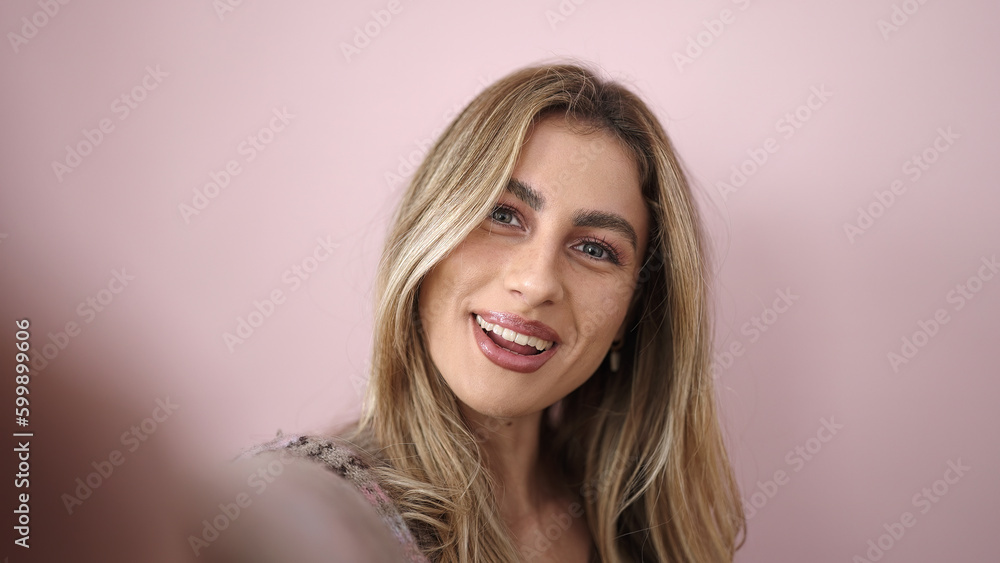 Fototapeta premium Young blonde woman smiling confident making selfie by camera over isolated pink background
