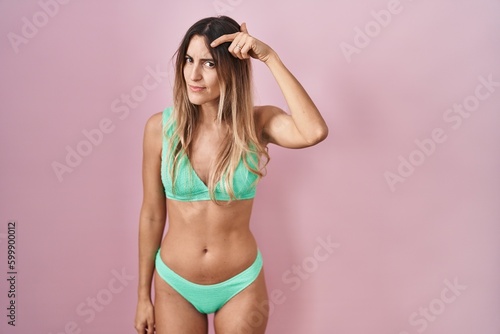 Young hispanic woman wearing bikini over pink background pointing unhappy to pimple on forehead, ugly infection of blackhead. acne and skin problem