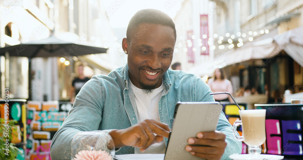 Good-looking smiling carefree modern african american with stylish beard and in round earrings browsing internet on i-pad while sitting in cozy street cafe in summertime Good-looking smiling carefree modern african american with stylish beard and in round earrings browsing internet on i-pad while sitting in cozy street cafe in summertime