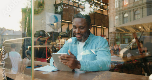 Good-looking smiling satisfied successful 30-aged black-skinned man in casual clothes sitting at the table in street cafe near the window and browsing internet on laptop